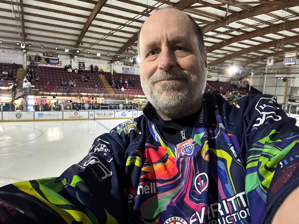 A man wearing a multi-coloured jersey, in front of an ice rink.