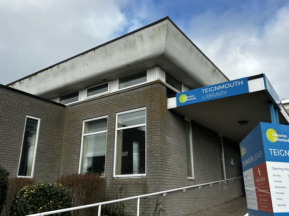 The entrance to a library. The building has a main block that rises to a row of clerestory windows and a white, curving soffit to a roof. To the left is a lower wing, and to the right is a long canopy and a sign with the opening hours. The walls are grey brick, with white window frames.