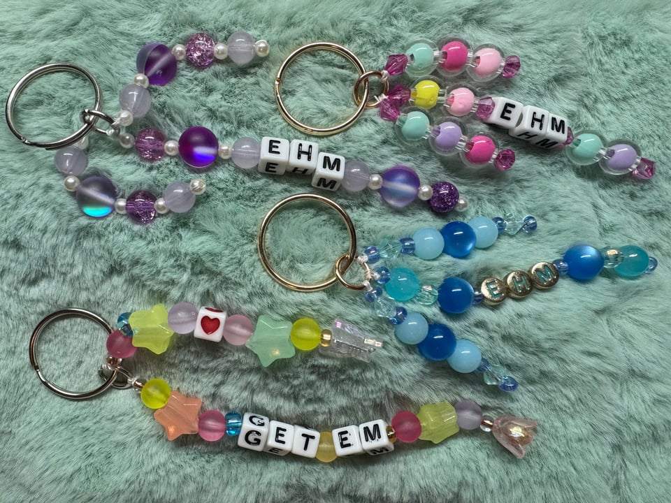A photograph of colorful keychains on a green fake fur background. The keychains say "EHM" and "GET EM".