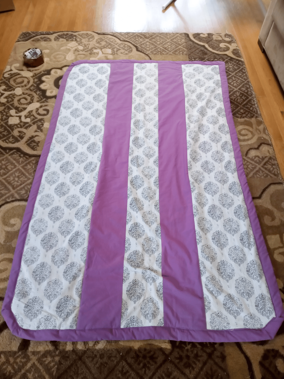 A quilt made of alternating stripes of purple and printed white fabric lays on a brown rug.