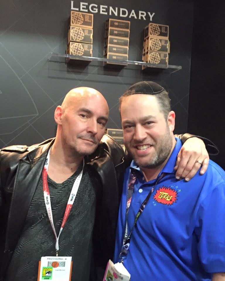 Grant Morrison wearing a black jacket and red and white badge lanyard with Neil Kleid wearing a blue topps star wars shirt and a black lanyard at the Legendary Comics booth