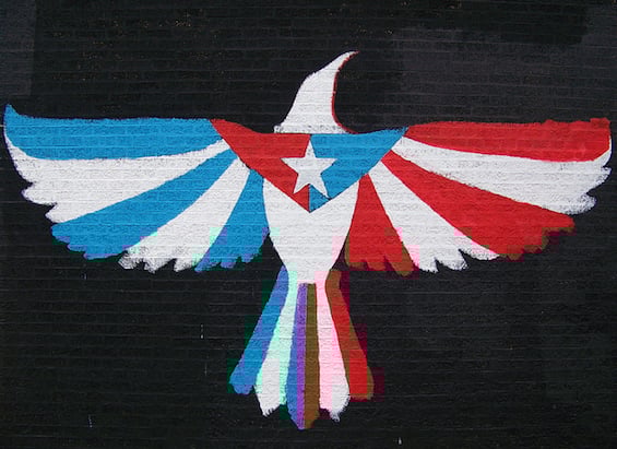 A mural: against a black brick wall, a white dove spreads its wings. The wing on the viewer's left is striped in light blue; the one on the viewer's right in red. The tailfeathers alternate, with a blue stripe to the left, a red to the right, and a double stripe in the center that is blue and red. On the bird's chest, a red triangle sits facing the blue stripes, a blue one facing the red stripes, and bisecting them is the white star that the Puerto Rican and Cuban flags share.