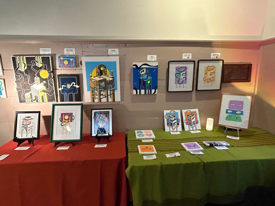 Paintings neatly arranged across the wall and tables. The artwork is unified by similar visual language (isometric views of architecture on stilts and/or limited color palettes).
