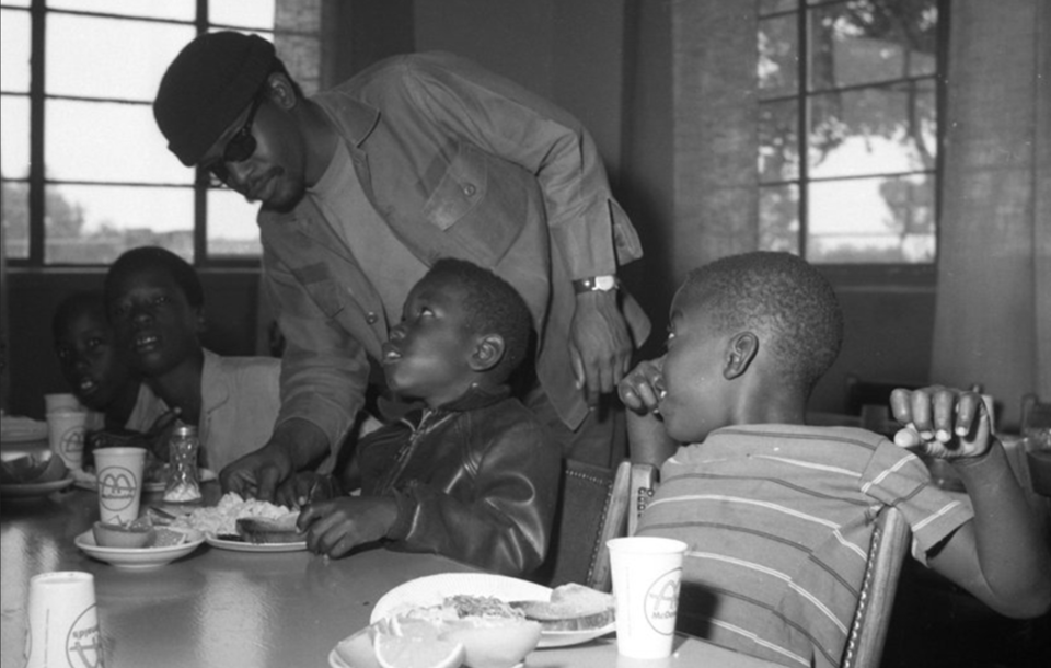 a young man with dark brown skin sunglasses and cap feed found young boys who look directly at the camera the young man or smile. the food includes drinks, sandwiches and orange slices all in a community hall with windows.