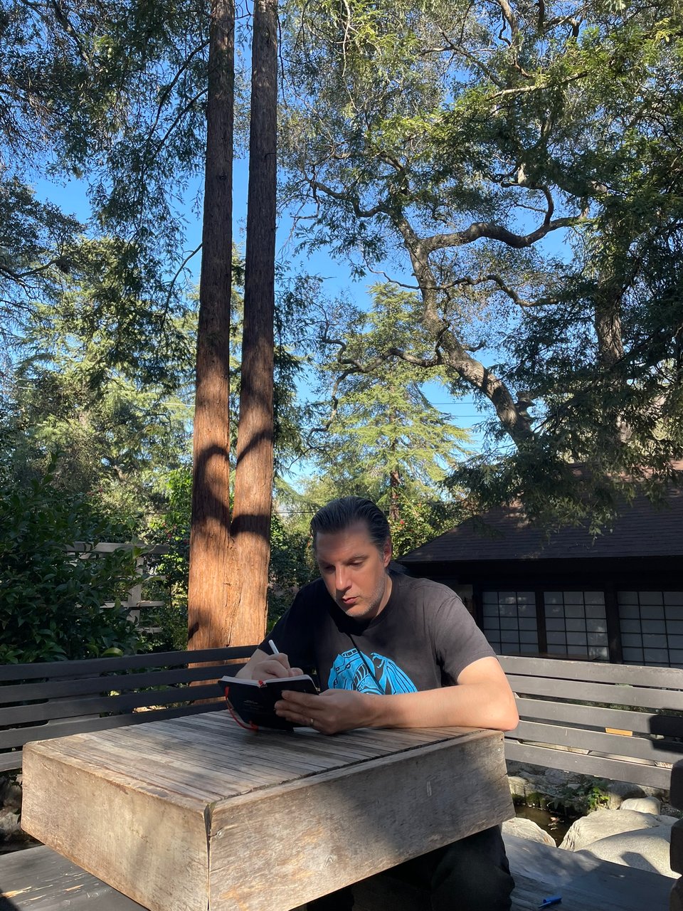 A man sits at a wooden table, surrounded by trees writing in a small notebook