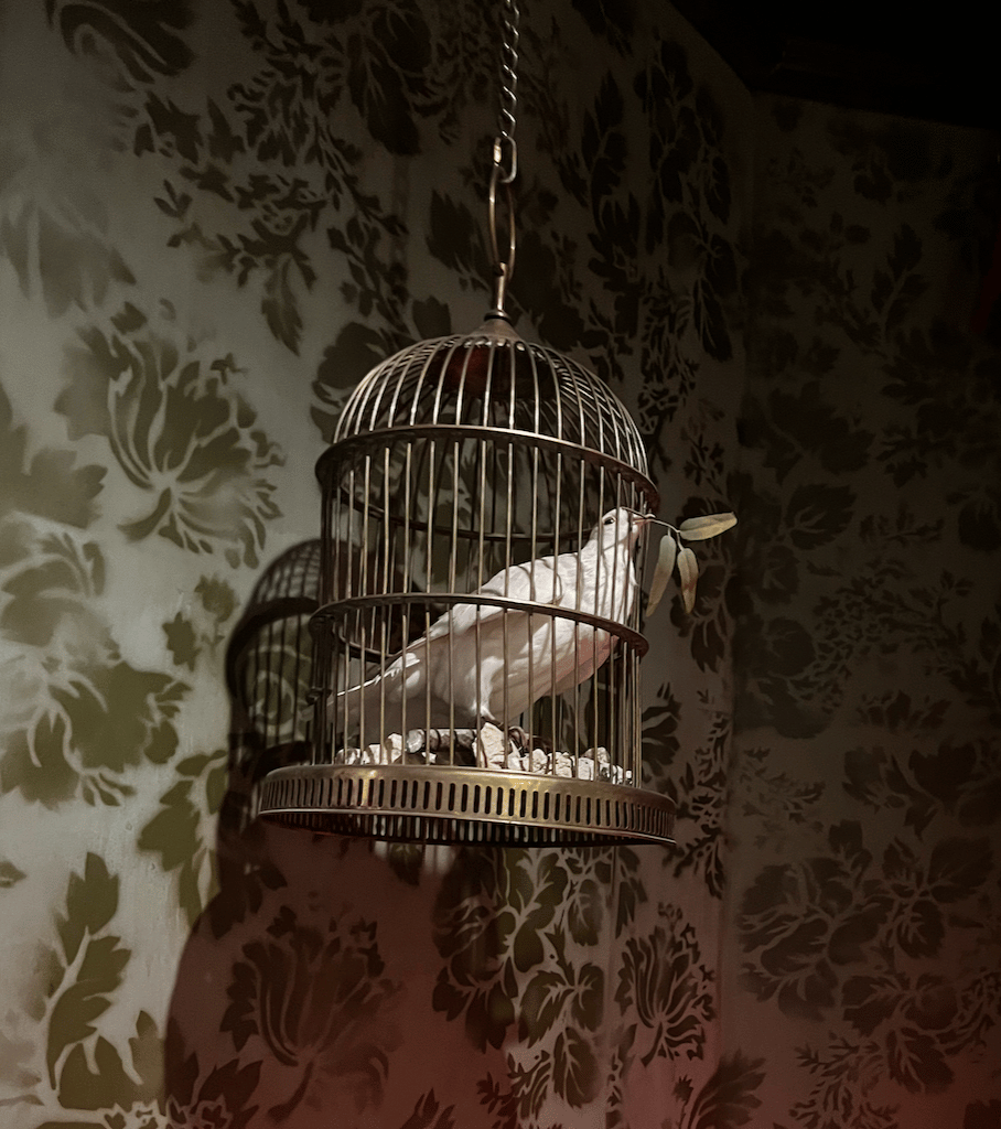 A white dove with an olive branch in its beak, inside a small cage. The cage is in front of flocked wallpaper
