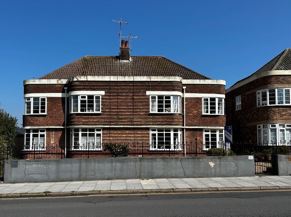 A 1930s brick apartment block in the sun. The front corners are curved, with windows. The main frontage uses bricks to create lines across it.