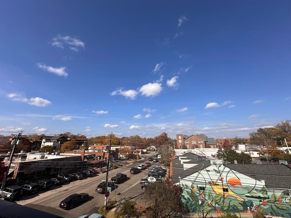 A wide angle photograph looking North by Northwest from the second floor terrace of the Drakeford Library Complex. The top three fifths of the image is a partly cloudy sky. At the horizon, just right of center, the Century Center can be seen with its "Black Lives Matter" mural. In the foreground at bottom right is a gigantic bicycle sculpture attached to the building that is home to Open Eye Cafe. A line of cars and trucks is stopped at a red light and extends from bottom left to the center of the frame. They are in the North bound lane of Greensboro.