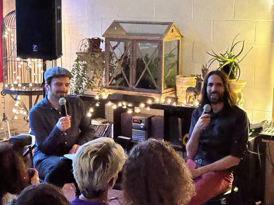 I am wearing a cap, seated to the left holding a microphone while Jerome, with long black hair, is seated at the right holding a microphone. Lights, plants, and an analogue sound system are against the wall behind us. The heads of the audience's front row are in the foreground.