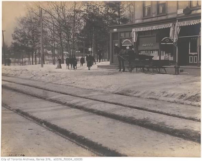 image showing a street with four clear streetcar track furrows in the foreground, and storefronts, pedestrians, and one horse-drawn sleigh in the background