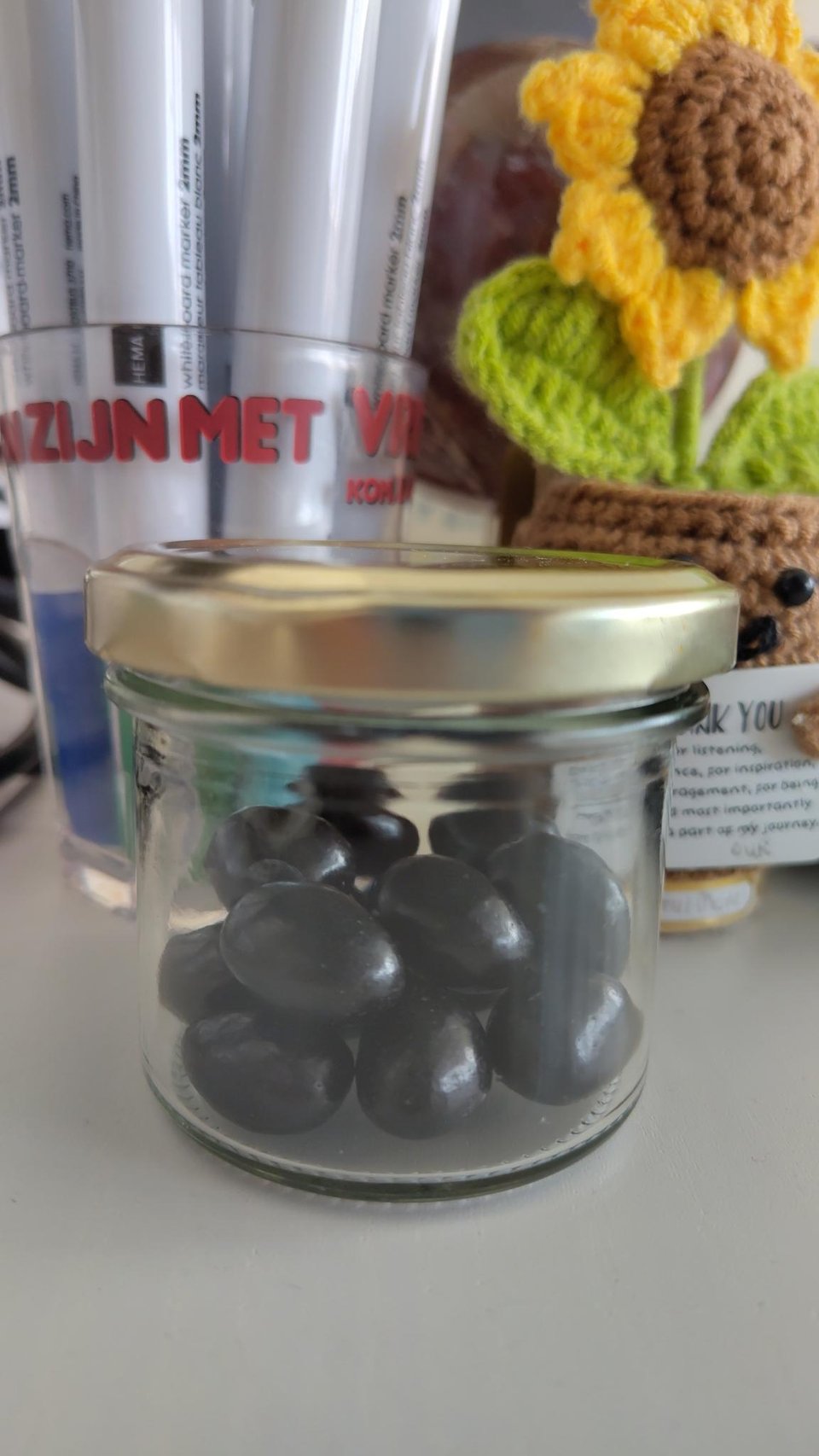 Image description: closeup of a small jar full of black jellybeans. it sits next to a clear plastic cup of dry erase markers and a small crocheted sunflower in a pot holding a thank-you note. End image description.