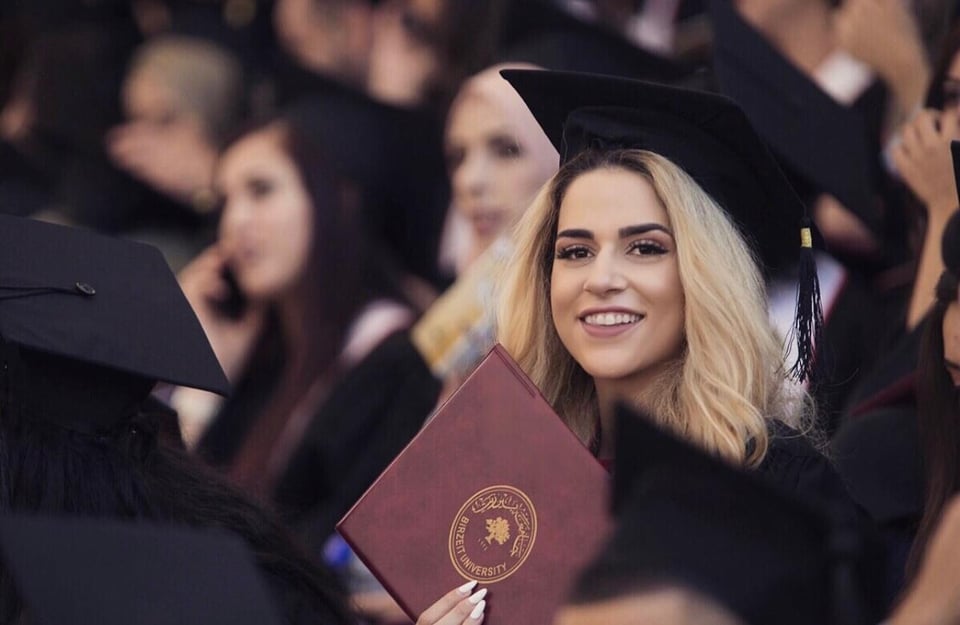 A woman with blonde hair in a graduation cap holding a diploma and smiling as she looks at the camera, among other people in graduation attire