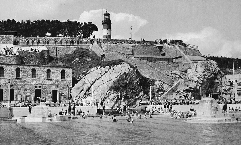 The semi-circular swimming pool with swimmers in it and fountains on. Walkways and stairs are cut into the cliffface. Where the current entrance building and changing block stands there is an old stone changing block that looks Victorian.