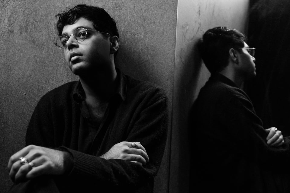 A black-and-white photo of man with glasses and short dark hair with his arms crossed, reflected in profile by a mirror to his left.