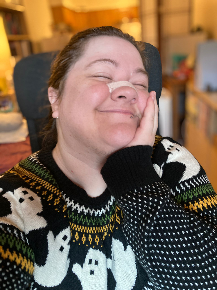 Jessi, a fat, white person with brown hair, smiling happily with a hand to their cheek and their eyes closed as if savoring the sensation of wearing their Fair Isle-style ghost sweater.