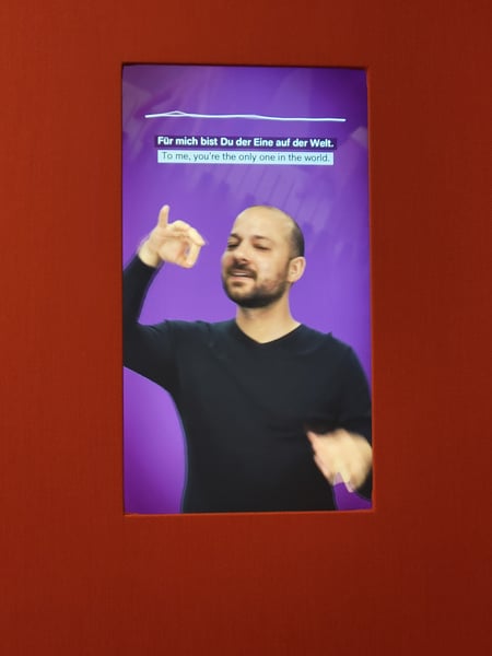 A video screen shows a man signing a song, with German and English subtitles.