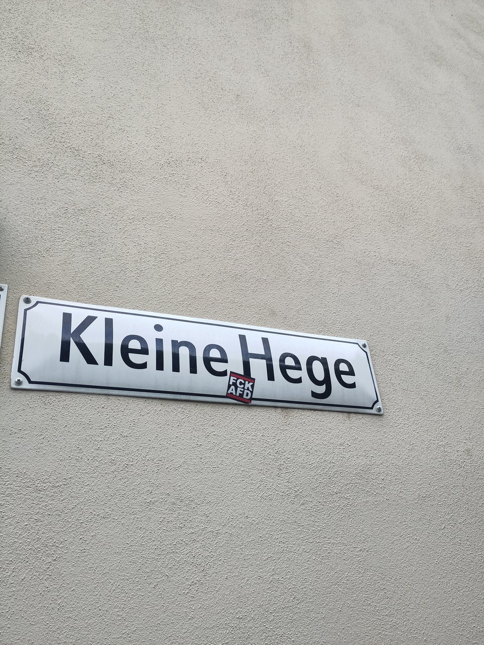 Grau verputzte Wand, daran ein Straßenschild mit der Aufschrift "Kleine Hege", darauf wiederum ein Aufkleber "FCK AFD"
