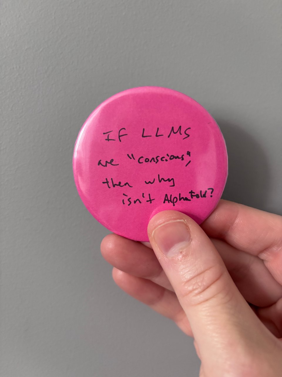 a light-skinned hand holds a pink button pin that says "If LLMs are 'conscious', then why isn't AlphaFold?" in black ink