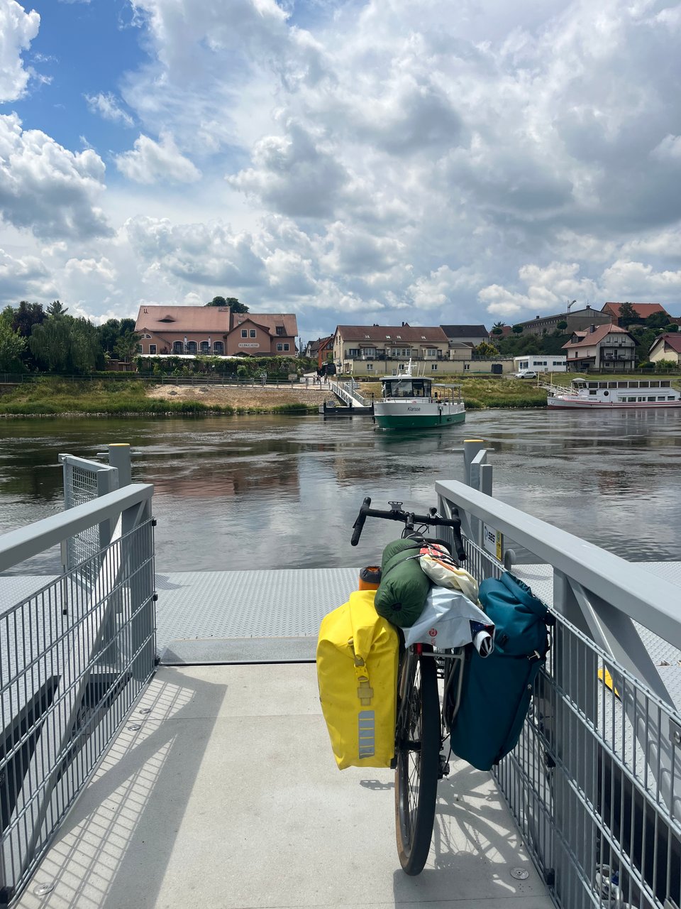 A heavily loaded bike on the gangway by a river, with a ferry halfway across.