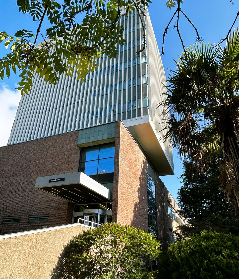 A pale six storey tower block resting on a mostly hidden base that connects it to a large brick mass with a vertical slot containing large windows and a deep cantilevered porch. The underside of the tower extends beyond the brick mass, and glows.