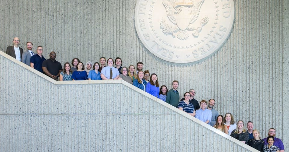 A large group of people poses for a photo on the steps inside a government building, with the large Seal of the United States prominently displayed above them. The individuals are arranged across multiple levels, smiling and looking toward the camera.