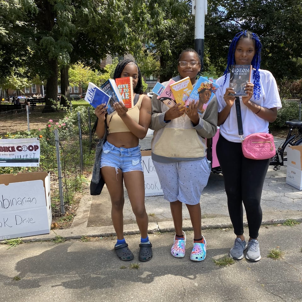 Three young, Black women hold up books in front of the donation box for the Nonbinarian Book Bike Book Drive