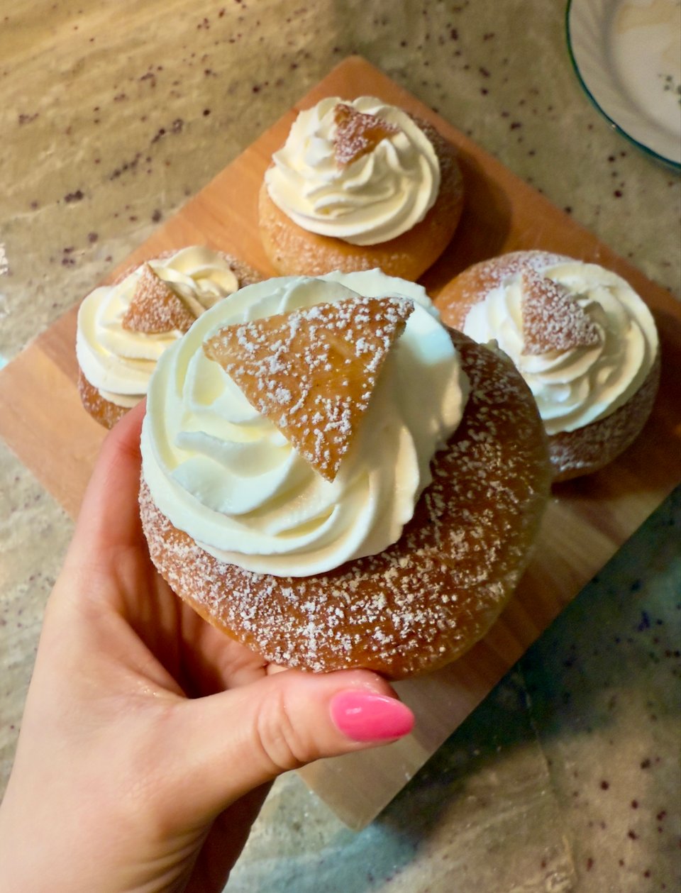 Four semlor buns, one held to camera by author, topped with cream and a triangle of dough and sprinkled with powdered sugar.