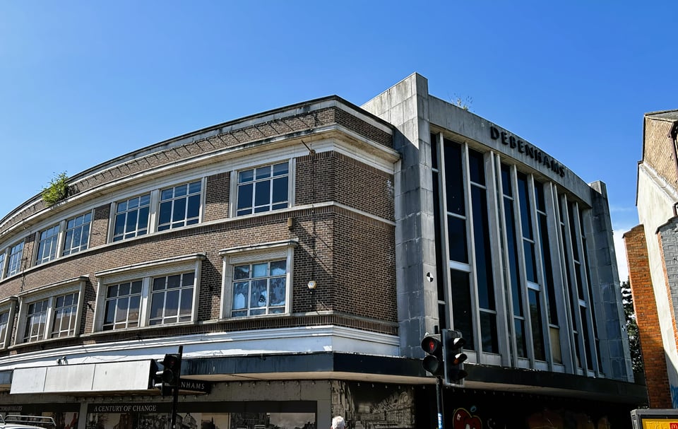 A corner of a boarded up Debenhams department store. At the front is a 1930s curved front of brick and stone with some light modern touches. At the side is a strongly vertical 1960s extension, with full height windows set into grey concrete frames.