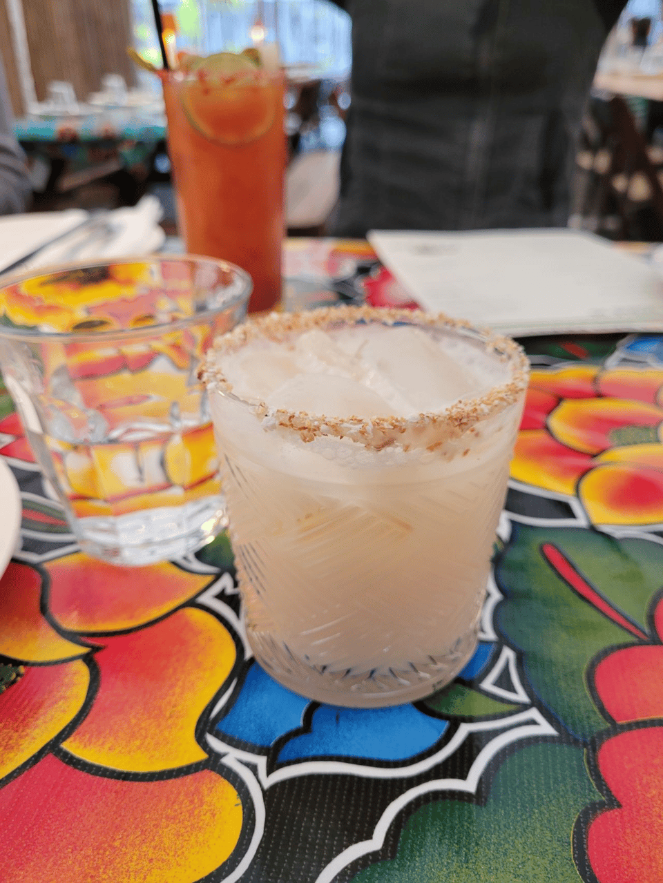 Three drinks are on a colorful floral tablecloth. In the background is a Bloody Mary, in the middle ground is a glass of water, and in the foreground is a coconut drink.