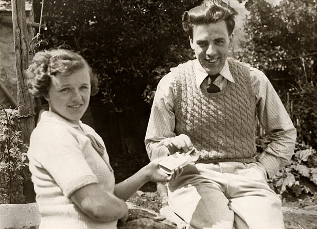 A black and white photograph of a woman (left) handing a man (right) a cigarette. The man is wearing a cable knit sweater vest and sitting on a wall.