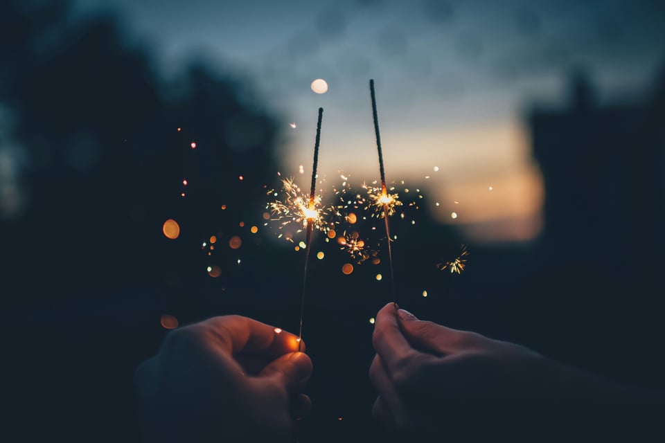Two hands holding sparklers in the foreground of a setting sun. Individual sparks are captured as they shoot off the sparkler stick.
