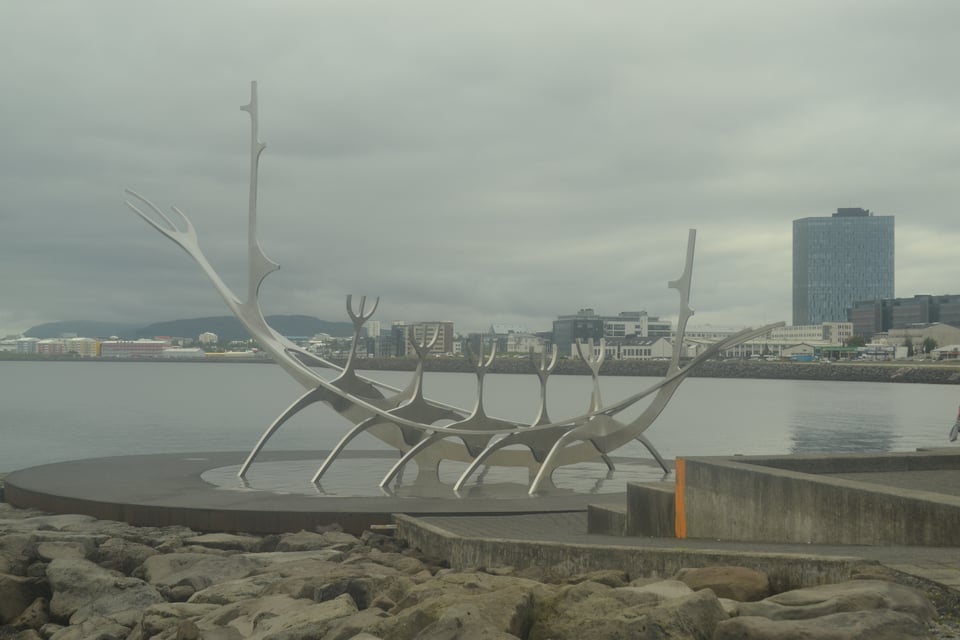 A sculpture that represents a ship with sailors rowing, made of silver metal. The sky is grey and it is in front of a bay, with buildings on the other side.