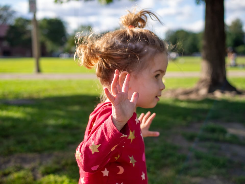 A profile portrait of Sona at the park, wearing a red pajama top with gold stars and moons. She is in motion, with her arms stretched out to the sides.