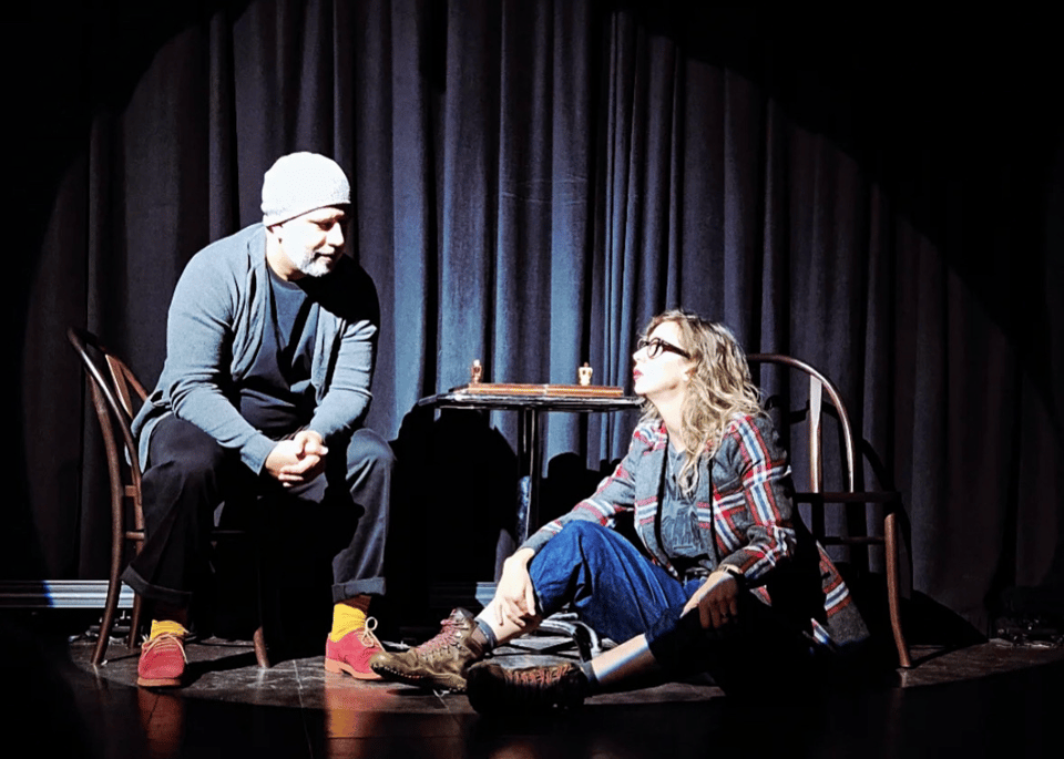 A man, seated in a chair, is speaking with a woman, sitting on the floor on a stage.