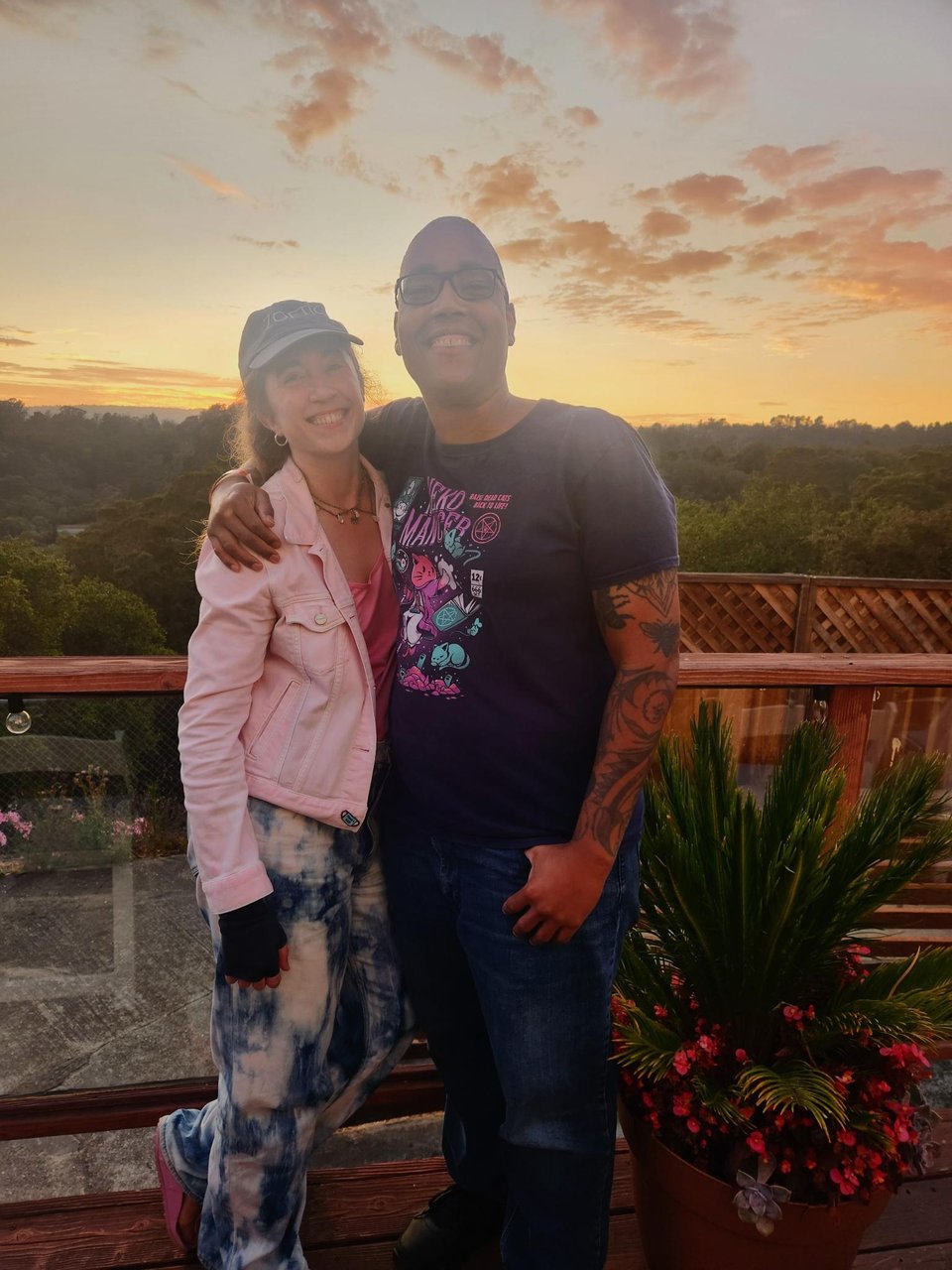 Hans and Saifa smiling, standing on a wooden balcony against a yellow-orange sunset in Santa Cruz. Saifa has an arm around Hans.