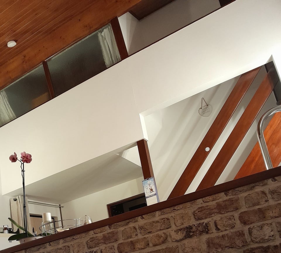 The back wall of the open plan living area, photographed from sitting down near the kitchen. In the foreground is a stone half-wall hiding the kitchen area. Beyond it, a simple wooden banister goes up. Above the front door is the master bedroom, with full width dappled glass windows. Running from the windows towards the camera is a wood-clad ceiling.