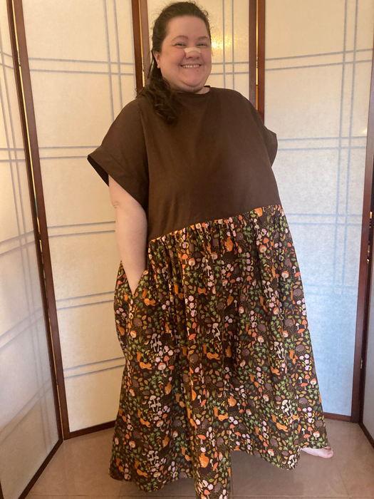Jessi, a fat, white person with wavy brown hair, stands in front of a divider wearing an oversized dress made with a dark brown short sleeve top attached to a brown, autumnal patterned skirt with illustrations of squirrels, mushrooms, hedgehogs, leaves, and so on. They look pleased with their work.
