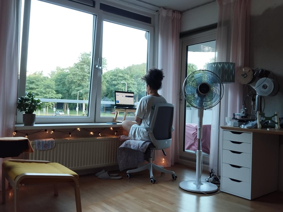 image description: a photo of me from behind; I’m seated with my legs criss-cross at a make-shift desk on a windowsill. I’m sitting in an office chair. There’s a standing fan near another desk behind me to my right. There’s a yellow footstool to my left. There are some fairy lights lining the bottom of the windowsill. Light shines in from the windows, and the treeline is visible outside. The windows are flanked by sheer, light pink curtains. End image description.