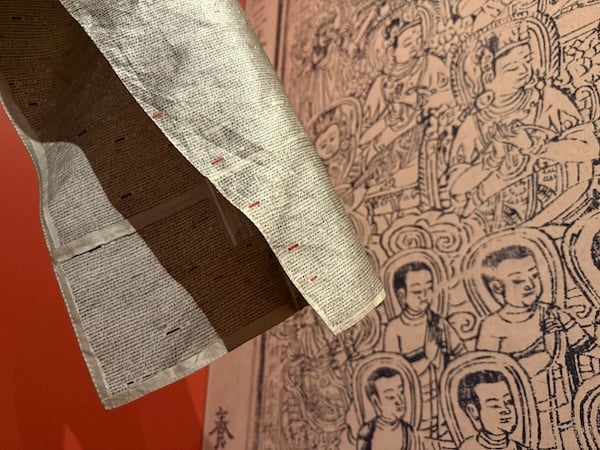 Sleeve of silver silk garment covered in tiny black Chinese characters against a background of multiple haloed Buddhist figures, photographed at the Princeton University Library exhibit Forms & Function: The Splendors of Global Book Making