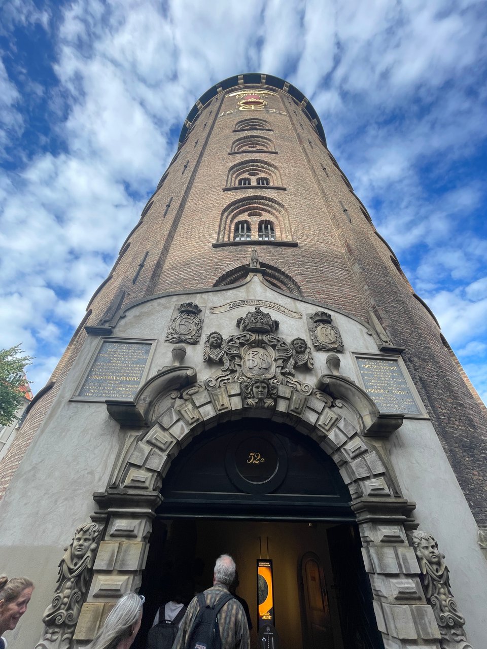 A man looks up at the entrance of a large brick towers with blue sky behind and spattered puffy clouds.