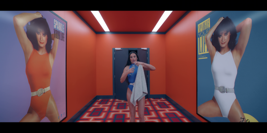 Demi Moore as Elisabeth Sparkle stands in the middle of an orange corridor with blue highlights and a carpet reminiscent of The Shining. She's wearing workout lycra and flanked on both sides by large promotional portraits of herself at an earlier age.