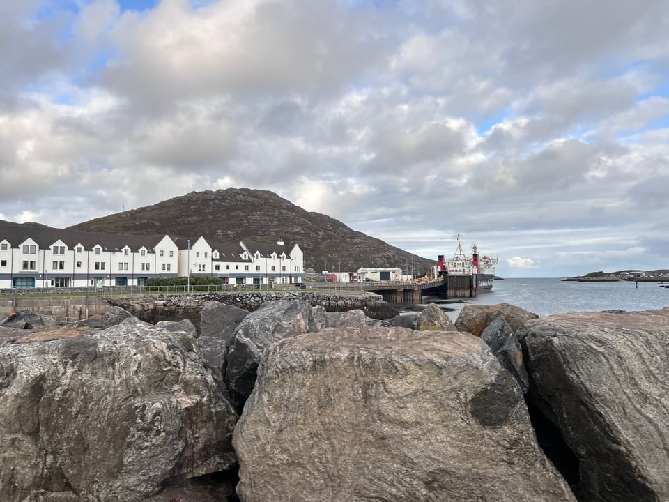 Lochboisdale harbour viewed from across the bay. A row of white shops stretches from the pier. A CalMac ferry waits.