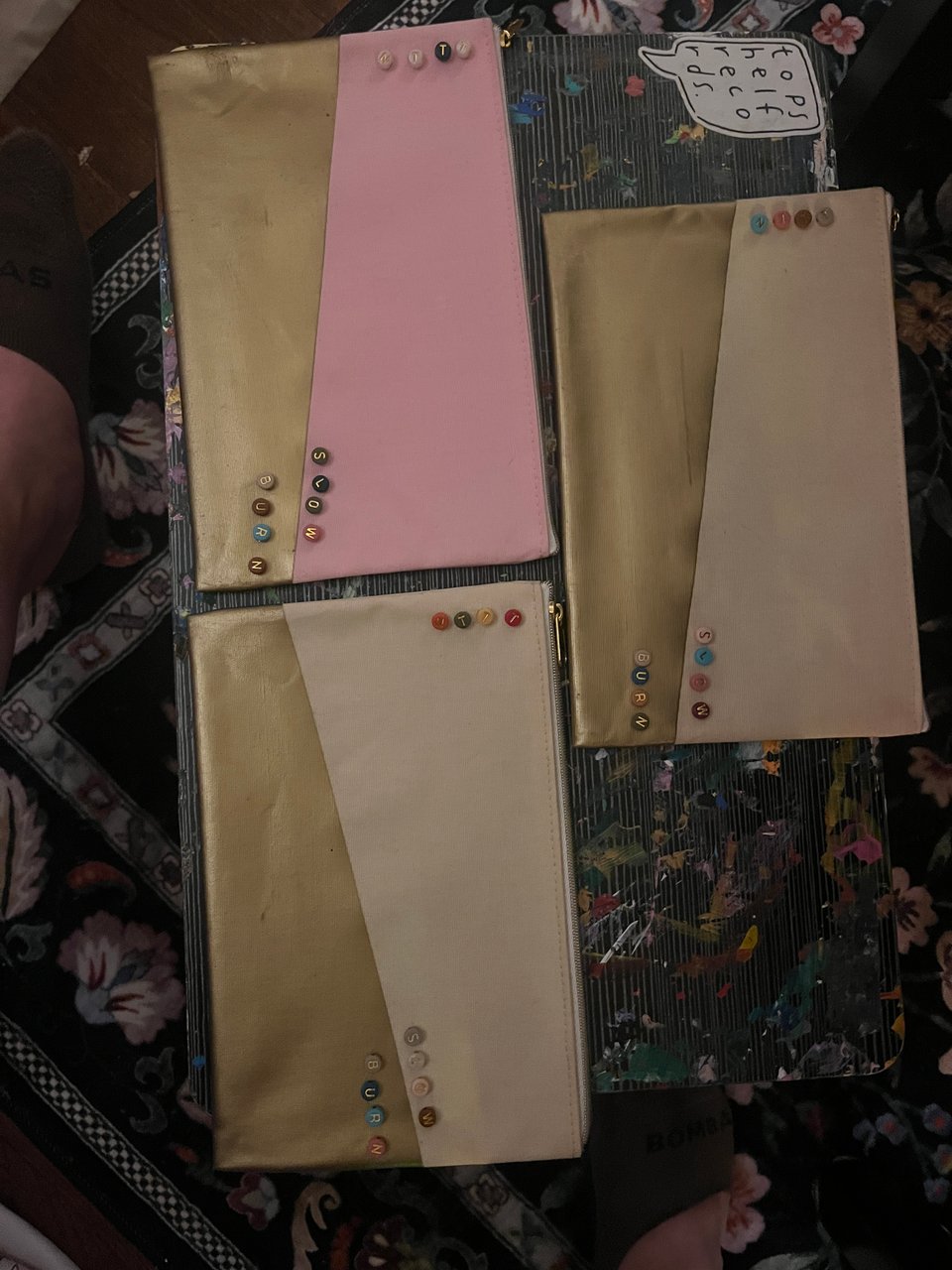Three pouches, one pink with gold and two beige with gold. All three have "Slow burn" and "NTTT" spelled out in round multicolored beads.