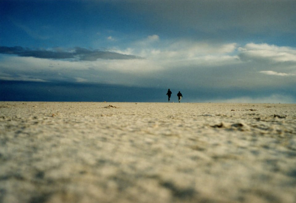 Photograph of the flat, salt flats landscape. Two figures can be seen in the near distance, running towards the horizon.