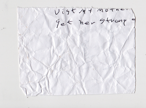 A crumpled square of paper. In the top right corner, in a thin and wiggly stroke, are two lines: "visit my mother" and "get her strong". The word "strong" is followed by an indistinct character -- possibly a dash -- that bleeds off the right-hand side of the page. The overall effect is mysterious