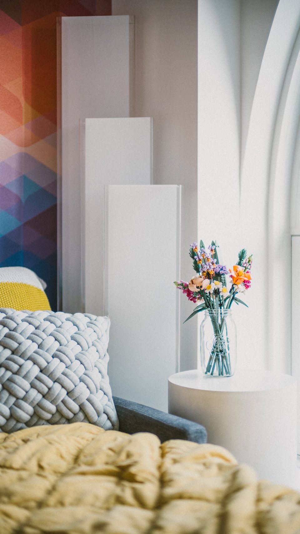 The corner of a bedroom with mutlicolored geometric wallpaper and a set of lego flowers on a side table.