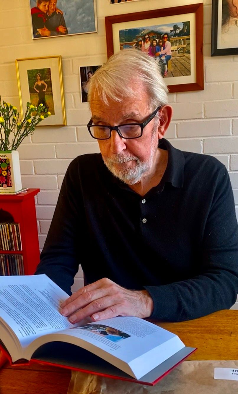 A man with white hair and glasses wearing a black shirt sits at a wooden table reading a large hardback copy of the book "Suddenly Something Clicked".