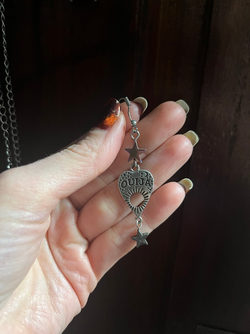 A woman's hand holding an earring that is an OUIJA board placard, with one star above it and one star hanging from it.