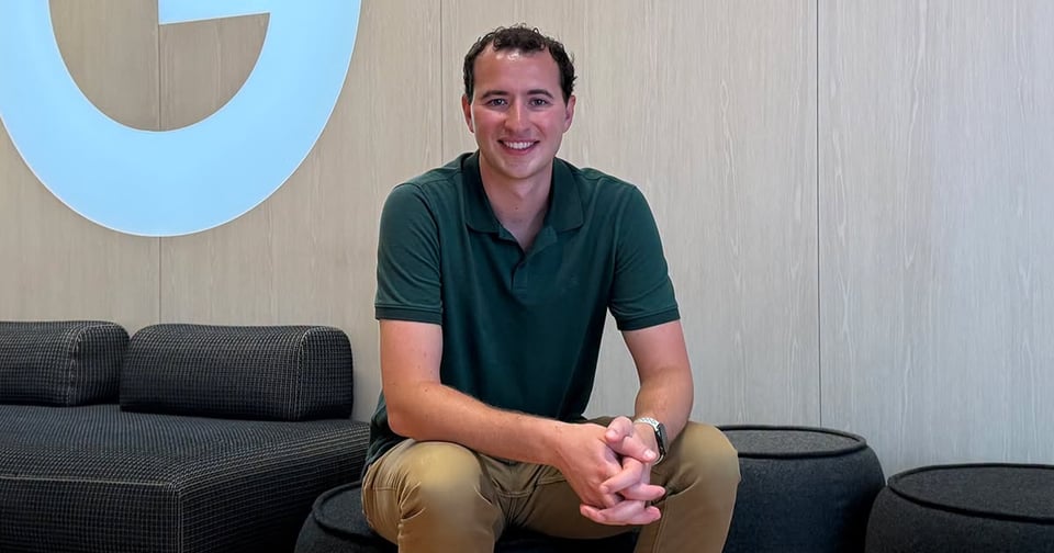 An adult with short dark hair sits on a black ottoman, smiling at the camera. He wears a green polo shirt and khaki pants, positioned in front of a light wood wall featuring a large blue circular logo.