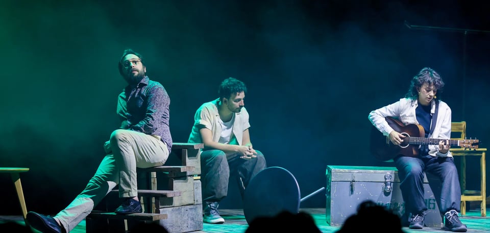 Two actors and one musician in the set of a theatre play, with furnitures and stair pieces cluttering the stage.